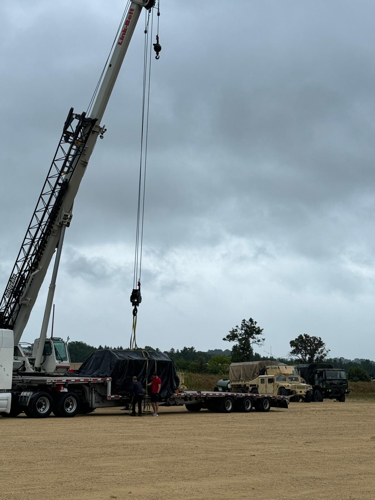 Crane rigging a covered load beside military vehicles for transport